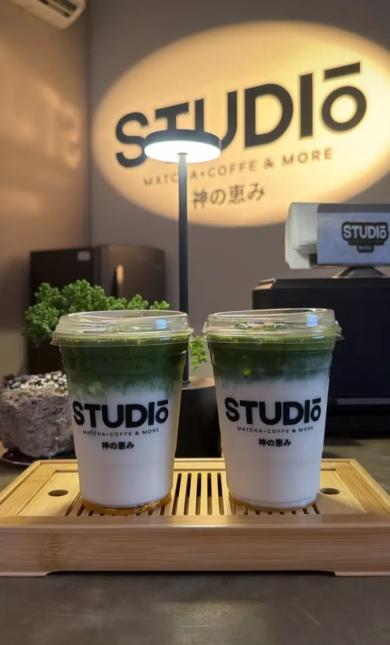 Dos matchas de Studio en vaso con su logo, sobre la charola de bambú, con el letrero encendido atrás